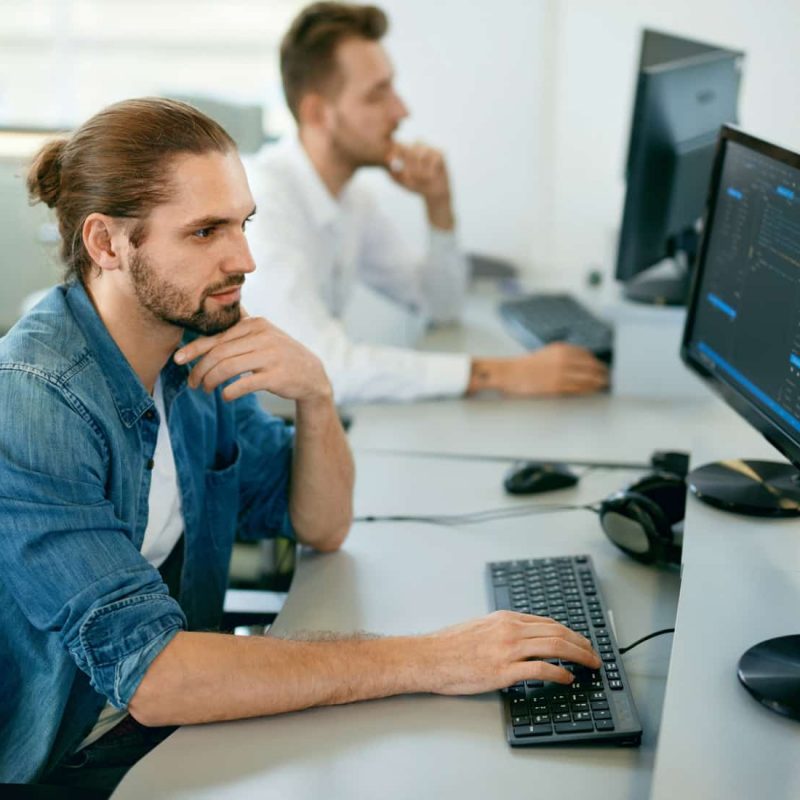 Programmers Working, Looking At Computer In IT Office. Handsome Young Men In Casual Closes Typing Codes, Working On Computer While Sitting At Workplace. High Quality Image.